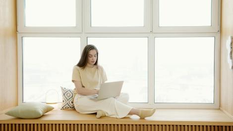 a woman sitting on a windowsill while using typing on her laptop